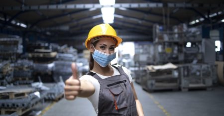 Woman standing in factory hall and showing thumbs up while wearing hygienic mask as prevention against corona virus.